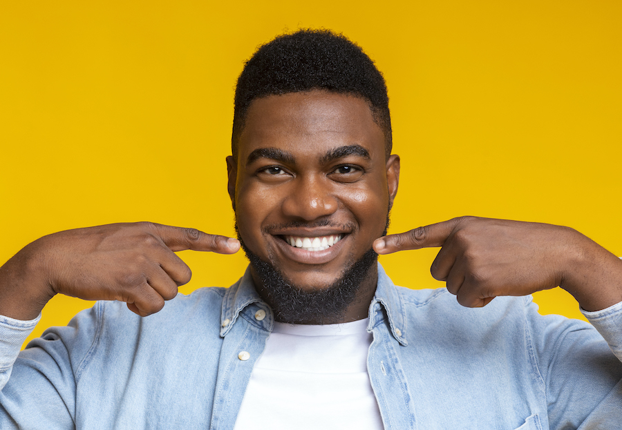 Black man smiles while pointing to his white teeth after professional teeth whitening at the dentist