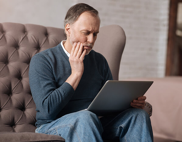 man holding his jaw in pain while looking at a laptop